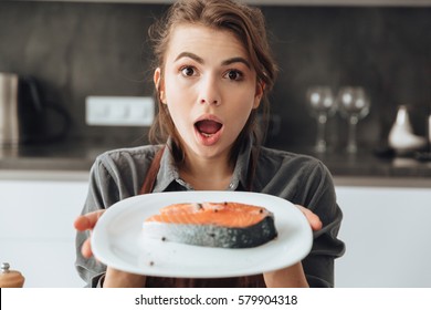 Picture Of Young Shocked Woman Standing In Kitchen While Cooking Fish. Looking At Camera.