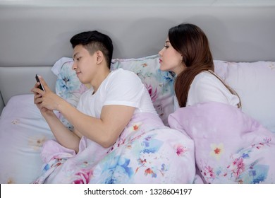 Picture Of Young Woman Snooping Phone Of Her Husband While Lying On The Bed