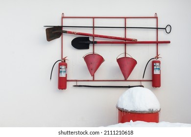 Pictured Is The Fire Safety Corner. Taken In Winter. White And Red Colors Predominate. In The Photo There Are Two Shovels, Fire Extinguishers, Cones, Crowbar, Hook, Red Barrel Of Sand