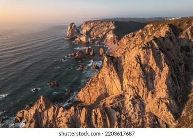 Picturesque Aerial View Of Rough Rocks And Ursa Beach Located On Shore Of Waving Sea Against Sundown Sky In Portugal