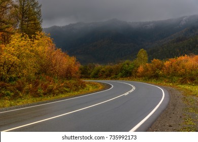 A picturesque autumn view of a steep turn of a wet asphalt road, mountains, golden trees and a dark stormy sky in cloudy weather