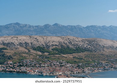 Picturesque coastal town with a mountain range backdrop, showing red-roofed houses, a calm bay, and rugged hills under a clear blue sky. Serene Mediterranean landscape captured from above.  - Powered by Shutterstock