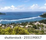 A picturesque coastal view with a clear blue ocean, waves crashing onto the shore, and lush green landscape. The foreground features buildings with beige roofs, palm trees, and vibrant vegetation.