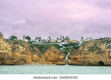 A picturesque coastal village atop rugged cliffs under a purple sunset sky. White stairways wind down the cliffs, Algarve, Portugal. - Powered by Shutterstock