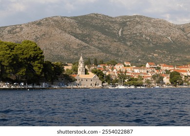 A picturesque coastal village features rustic buildings, a stone church with a tower, and lush trees lining the shore, set against a stunning mountainous landscape under clear skies. - Powered by Shutterstock