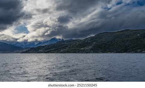 The picturesque coastline. Snow-capped mountain peaks of the Andes against a blue sky, clouds. Green vegetation on the coastal hills. The city buildings of Ushuaia on the shore. Ripples on the ocean - Powered by Shutterstock