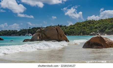 Picturesque Granite Boulders In The Surf On The Beach Of A Tropical Island. The Waves Beat Against The Rocks, Foam And Spread On The Sand. Splashes In The Air. A Green Hill Against A Blue Sky Clouds