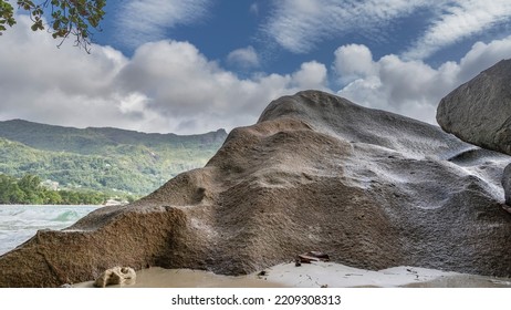 A Picturesque Granite Huge Boulder On A Sandy Tropical Beach. Close-up. A Wave Of Turquoise Ocean, A Green Hill Is Visible In The Distance. Blue Sky, Clouds. Seychelles. Mahe