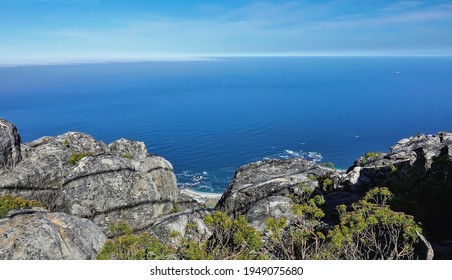 Picturesque gray spotted boulders against the backdrop of the blue Atlantic Ocean. A section of the coastline is visible. Fynbos bushes grow on the stones. Table Mountain top. Cape Town. South Africa 