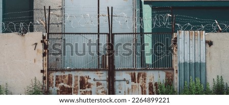 Picturesque industrial building close-up. Metallic rusty gate and concrete fence with barbed wire. Entrance in old factory. Closed area. Old industrial object is overgrown with grass. High metal doors