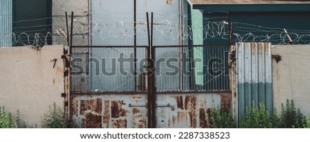 Picturesque industrial building close-up. Metallic rusty gate and concrete fence with barbed wire. Entrance in old factory. Closed area. Old industrial object is overgrown with grass. High metal doors