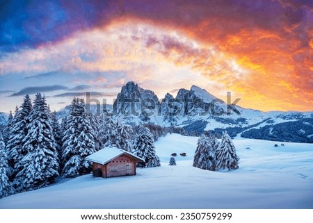 Picturesque landscape with small wooden log cabin on meadow Alpe di Siusi on sunset time. Seiser Alm, Dolomites, Italy. Snowy hills with orange larch and Sassolungo and Langkofel mountains group