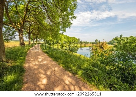 Picturesque landscape with trees and a curving sandy path along a lake. The photo was taken in the Dutch province of North Brabant on a partly cloudy day in the spring season.