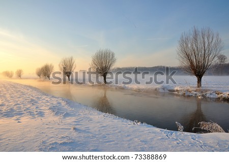 Picturesque and misty Dutch winter landscape with low sun, snow and partially ice on a narrow stream. Along the waterfront are bare willows. It is an award-winning photo of the municipality Drimmelen.