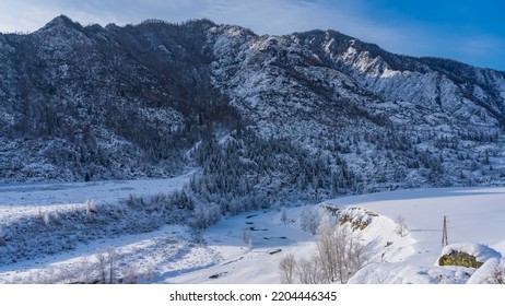 A Picturesque Mountain Against The Blue Sky. The Frozen River Winds Through A Valley Covered With Pure Untouched White Snow. Frosted Trees On The Banks. Towers Of Power Lines Are Visible. Altai