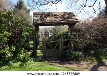 The picturesque old lychgate frames the tiny old Saxon church in spring sunshine at Duntisbourne Rouse in the Cotswolds, Gloucestershire, UK