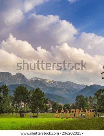 The picturesque photograph captures Zakopane, Poland, framed by the majestic Tatra Mountains under a cloudy sky. The city sign stands tall on the lush field of the city park. Travel concept