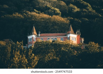 A picturesque pink historical villa with pointed rooftops nestled among lush green trees at sunset in Sintra, Portugal. The building's charming architecture contrasts with the forested hillside - Powered by Shutterstock