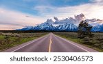 A picturesque road stretches towards the majestic, snow-capped Grand Teton Mountains under a vibrant sky. The landscape captures the serene beauty of Wyoming, evoking feelings of adventure