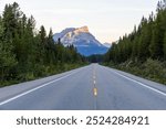 A picturesque road stretches towards a stunning mountain in Banff, Alberta, Canada, bordered by a dense forest. Experience the tranquility and beauty of nature in this breathtaking landscape.