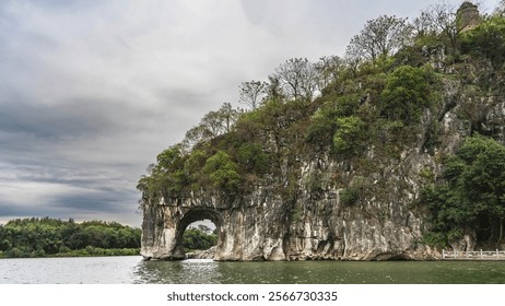 A picturesque rock by the river bank. Round arched opening above the water. Green vegetation on steep rocky slopes. A stone tower against a cloudy sky. China. Elephant Trunk Hill. Xiangbishan. Guilin. - Powered by Shutterstock