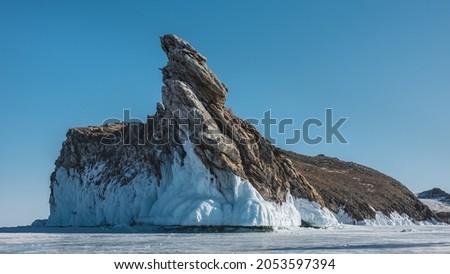 A picturesque rock, devoid of vegetation, rises on a frozen lake against the blue sky. Bizarre shapes, cracks on the stones. The base is covered with a thick layer of icicles. Baikal. 