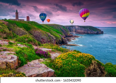 Picturesque rocky coastline with beautiful flowers and colorful hot air balloons over the sea at sunset. Fantastic lighthouse and stunning landscape, Cap Frehel, Brittany, France, Europe