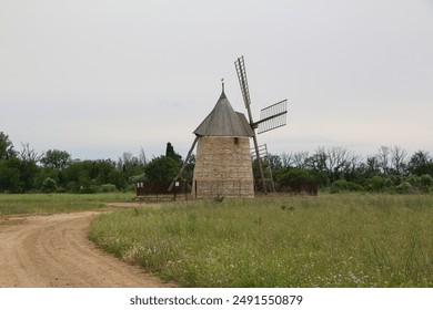 A picturesque rural landscape featuring an old stone windmill surrounded by a vast green field and a winding dirt path. The historic structure, set against a backdrop of lush trees and an overcast sky - Powered by Shutterstock