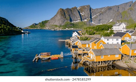 A picturesque scene of colorful wooden cabins perched on stilts over the clear blue waters of a Norwegian fjord, surrounded by lush green hills and majestic mountains. Sakrisoy, Lofoten, Norway - Powered by Shutterstock