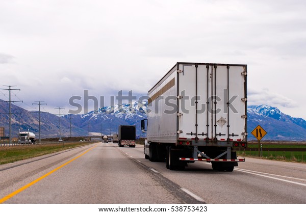Picturesque straight\
multilane highway with a dividing strip oncoming traffic flows with\
a semi trucks and trailers and snow-capped mountains of Utah in the\
clouds on the\
horizon.