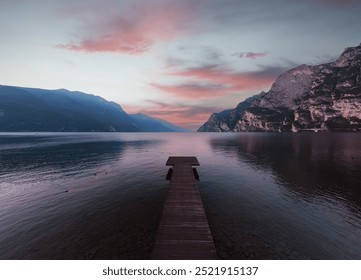 A picturesque sunset over a lake in Riva del Garda, with a wooden pier in the foreground and majestic mountains in the background. Calm waters and a colorful sky create an amazing view. - Powered by Shutterstock