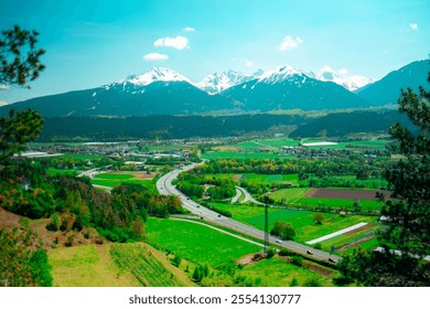 Picturesque valley features lush green fields, winding roads, and majestic snow-capped mountains under a clear blue sky, capturing the serene beauty of a vibrant spring landscape. - Powered by Shutterstock