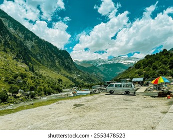 A picturesque valley scene showcasing lush green mountains, snow-capped peaks, a flowing river, and a small roadside market - Powered by Shutterstock