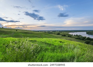 A picturesque view of a grassy hillside overlooking a meandering river in the distance. The sky is a vibrant blue with fluffy white clouds, creating a peaceful and tranquil atmosphere. - Powered by Shutterstock