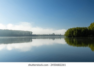 Picturesque view of a river shrouded in fog with the calm water reflecting the faint colors of the sunrise. The details of the trees in the background hidden by the fog add mystery to the scene.