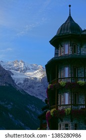 Picturesque View Of The Snowy Mountain Range Behind A Lovely Rustic Hotel In Dolomites. Beautiful Shot Of Colorful Flowers Covering The Balcony Of A Stone Hotel High In The Tranquil Italian Mountains.