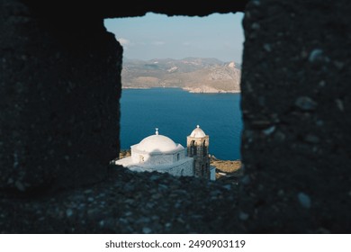 A picturesque view of a white-domed church overlooking a deep blue sea, framed through a stone window, with mountains in the background, capturing the serene beauty of the coastal landscape. - Powered by Shutterstock