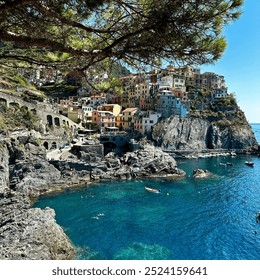 The picturesque village of Manarola features vibrant houses perched on steep cliffs, surrounded by azure water. Visitors enjoy the serene beach and rocky shoreline under bright sunlight. - Powered by Shutterstock