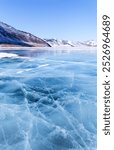 Picturesque winter landscape of frozen Baikal Lake on cold sunny day. Shallow bay is covered with blue ice with cracks against backdrop of snowy coastal mountains. Beautiful natural winter background