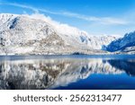 Picturesque winter landscape with tranquil lake mirroring snow-capped mountains under a vibrant blue sky. Panoramic nature scenic view