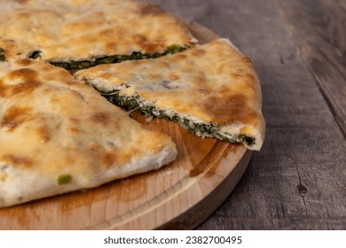 Pie with spinach, herbs and suluguni cheese on a wooden board. Whole pie with cut off piece. Traditional round flat pie with filling. Homemade pastries of Caucasian and Balkarian cuisine. - Shutterstock ID 2382700495