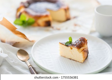 A Piece Of Basque Brunt Cheese Cake Is Put On A White Plate With Mint On Top As A Whole Cake Is In The Background Horizontal