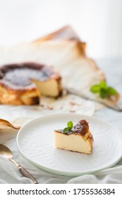 A Piece Of Basque Brunt Cheese Cake Is Put On A White Plate With Mint On Top As A Whole Cake Is In The Background Vertical