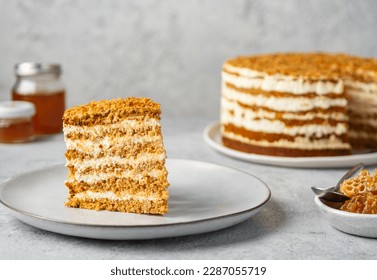 Piece of biscuit cake Medovik. Layered caramel honey cake with whipped cream and sour cream. Honeycomb and jars of honey on concrete background.
