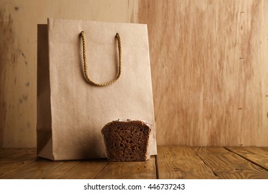 Piece Of Brown Rye Bread Presented Near Take Away Blank Bag From Craft Paper In Artisan Bakery On Wooden Background