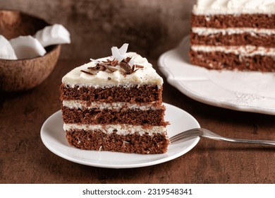 A piece of chocolate cake with fresh coconut cream on a dark brown background. Coconut fruit on background. - Powered by Shutterstock