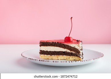 Piece Of Chocolate Vanilla Cake With Maraschino Cherry. Side View Cake On White Plate On Pink Background