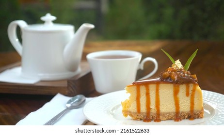 Piece Of Delicious Fresh Cheesecake Cake On Wooden Table With White Teapot In Outdoor Cafe. Tasty Fresh Pastry Decorated With Edible Gold And Caramel With Cup Of Hot Tea. English Tea Time. Sweet Food