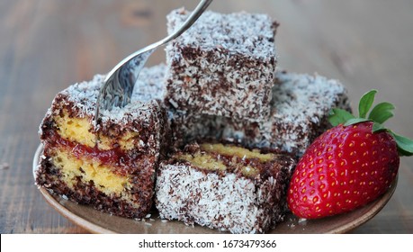 Piece of lamington cake with strawberry jam is picked up with a fork. Squares of sponge cake coated in chocolate sauce and rolled in desiccated coconut on plate.