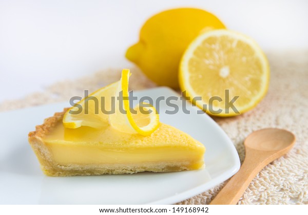 a piece of lemon tart on a white plate\
decorated with lemons and wooden\
spoon